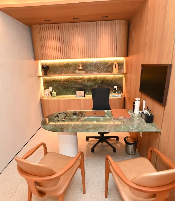 Consultório do INME com mesa de mármore verde, duas poltronas caramelo e televisão na parede